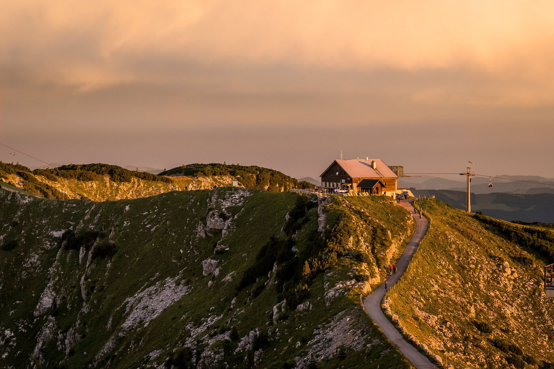 Der Sonnenuntergang taucht die majestätischen Berge in ein warmes, goldenes Licht und schafft eine friedliche Atmosphäre. Die sanften Hügel und die charmante Hütte laden dazu ein, die Schönheit der Natur zu genießen und den Tag in Ruhe ausklingen zu lassen.