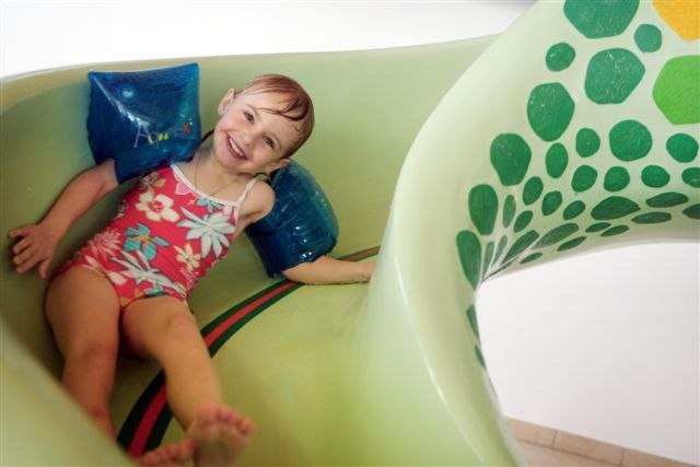 Ein lachendes Kind mit Schwimmflügeln rutscht eine grüne Wasserrutsche hinunter.