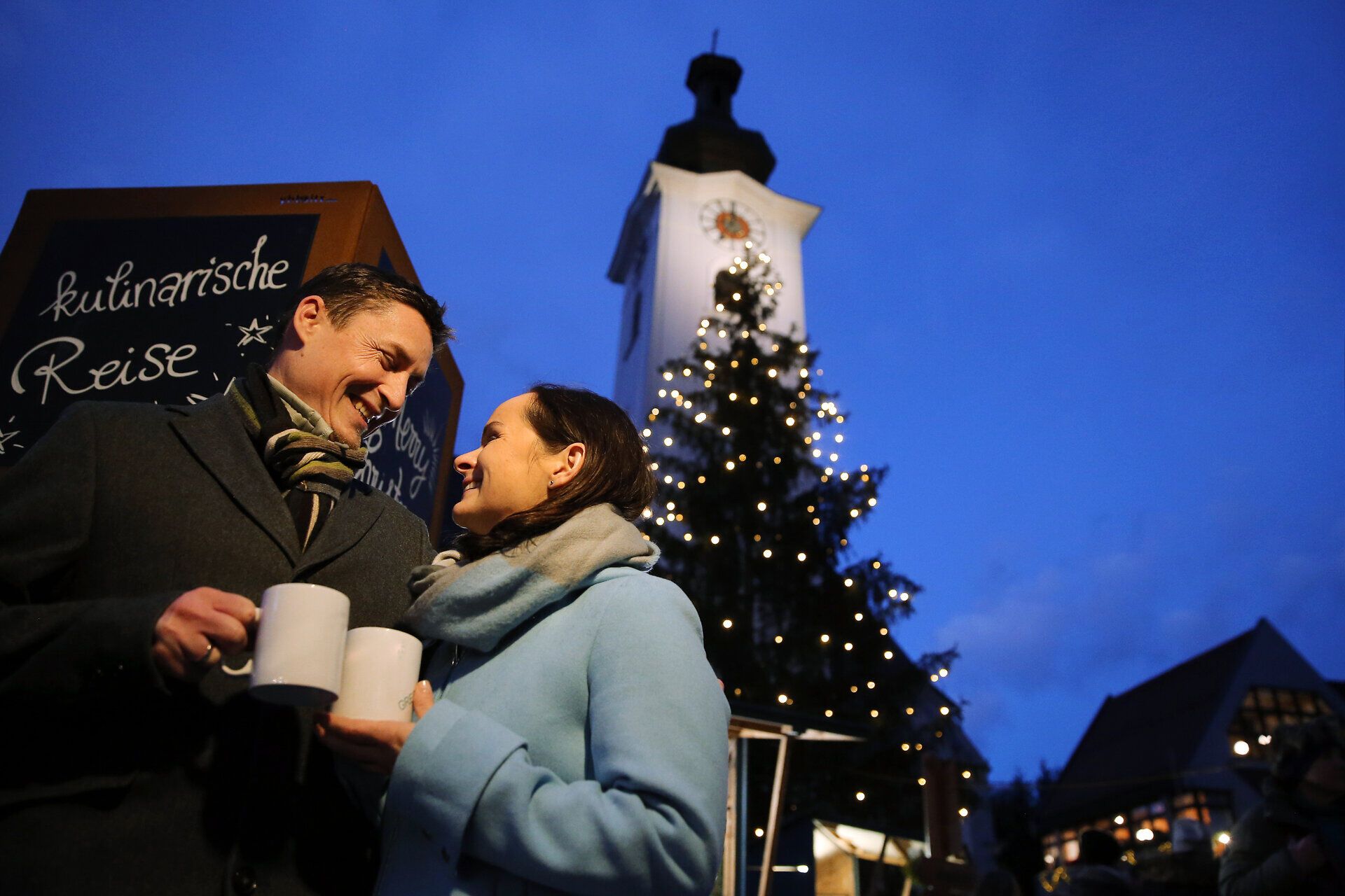In der kalten Abendluft strahlen die Lichter des Adventmarktes und schaffen eine festliche Stimmung. Paare genießen heiße Getränke und die romantische Atmosphäre unter dem funkelnden Weihnachtsbaum. Der Duft von Glühwein und frisch gebackenen Leckereien erfüllt die Luft und lädt zum Verweilen ein.