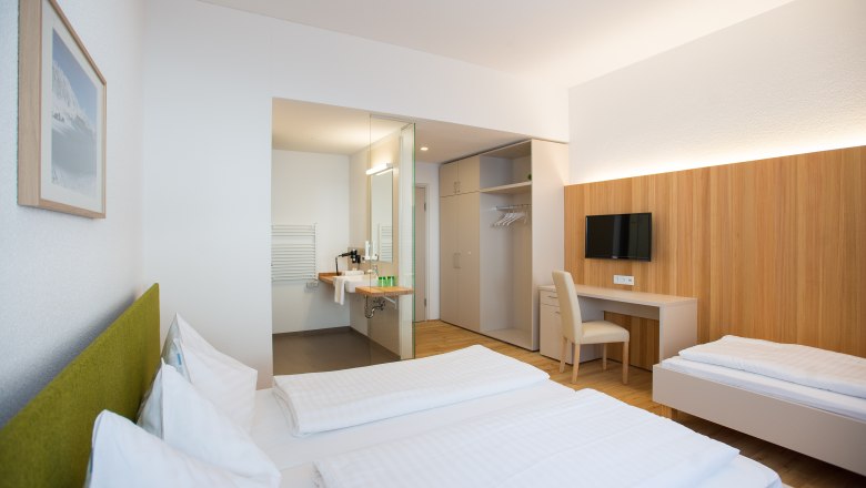 Modern eingerichtetes Hotelzimmer mit zwei Betten, Schreibtisch, Fernseher und offenem Badezimmer.