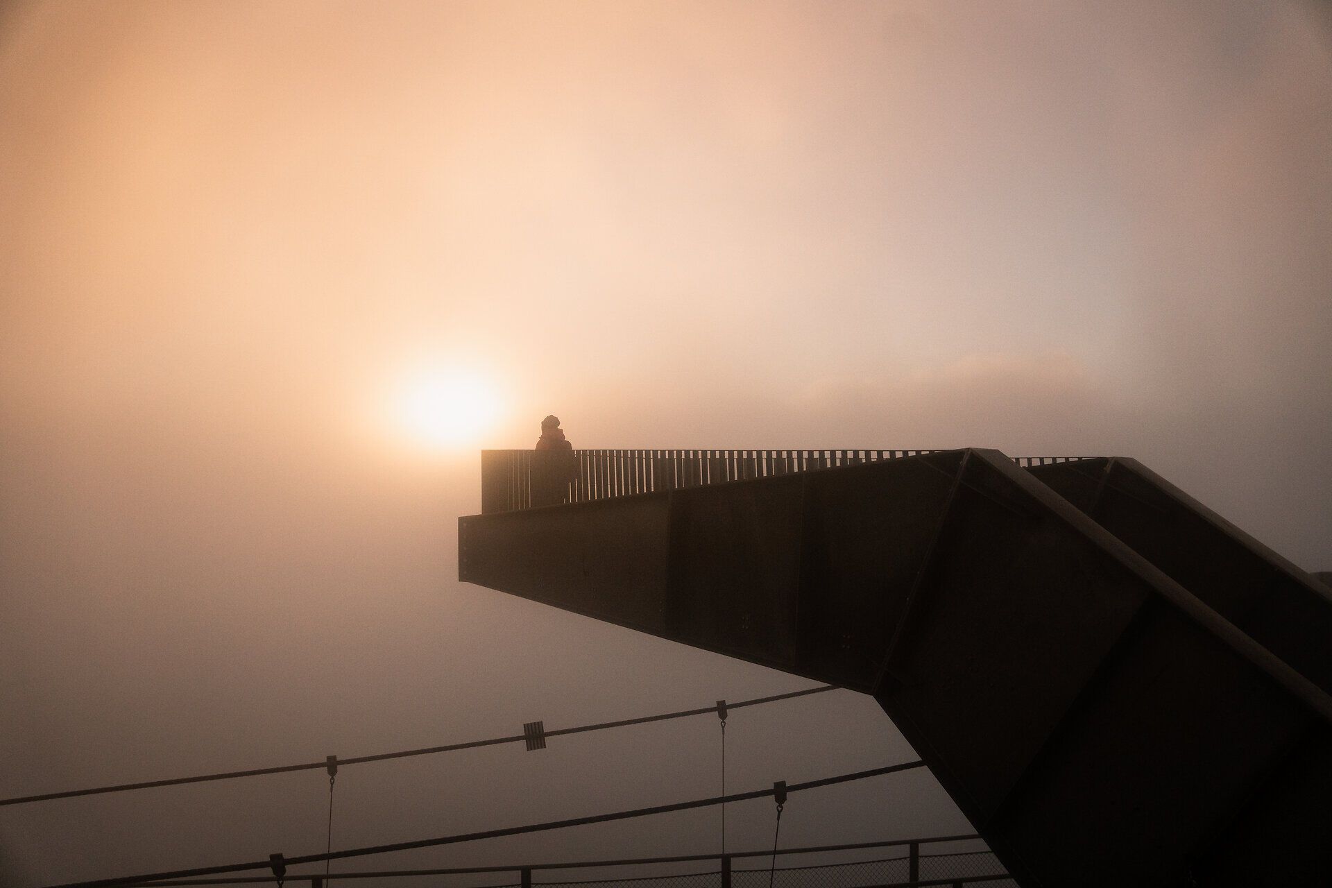 In der sanften Umarmung des Morgens, umhüllt von mystischem Nebel, steht eine Person auf der Aussichtsplattform und genießt den Anblick der aufgehenden Sonne. Die ruhige Atmosphäre und die majestätischen Berge laden dazu ein, die Seele baumeln zu lassen und die Schönheit der Natur zu erleben.